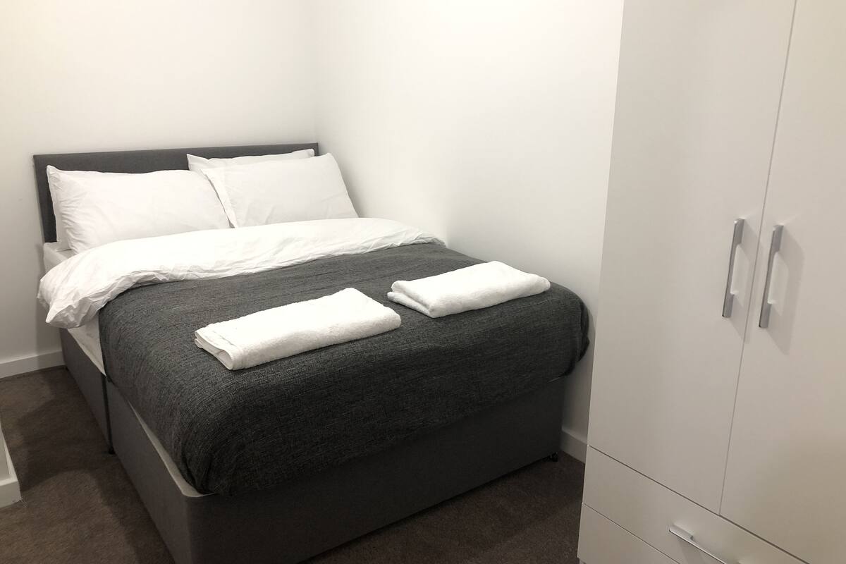 Una cama bien hecha con sábanas blancas y una manta gris, un armario con pomos plateados y suelo cubierto de alfombra.
