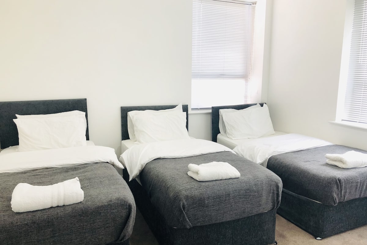 Habitación de hotel con tres camas individuales, cada una con cabecero gris y ropa de cama blanca.