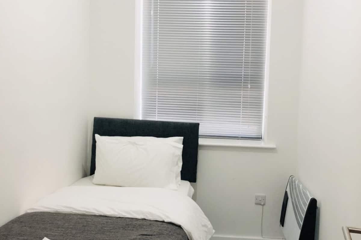 Una habitación pequeña y limpia con cama, ventana con persianas, radiador e iluminación.