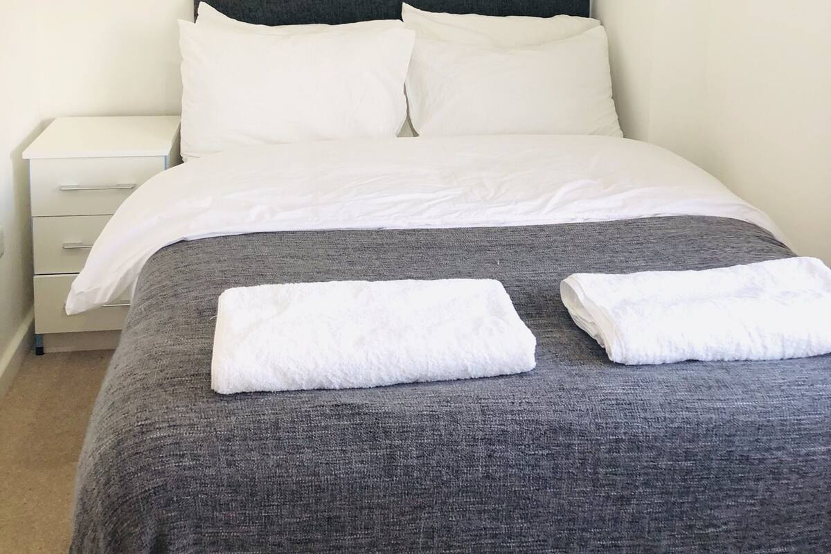 Una cama bien hecha con sábanas blancas y una manta gris, una mesita de noche blanca y un cabecero gris.