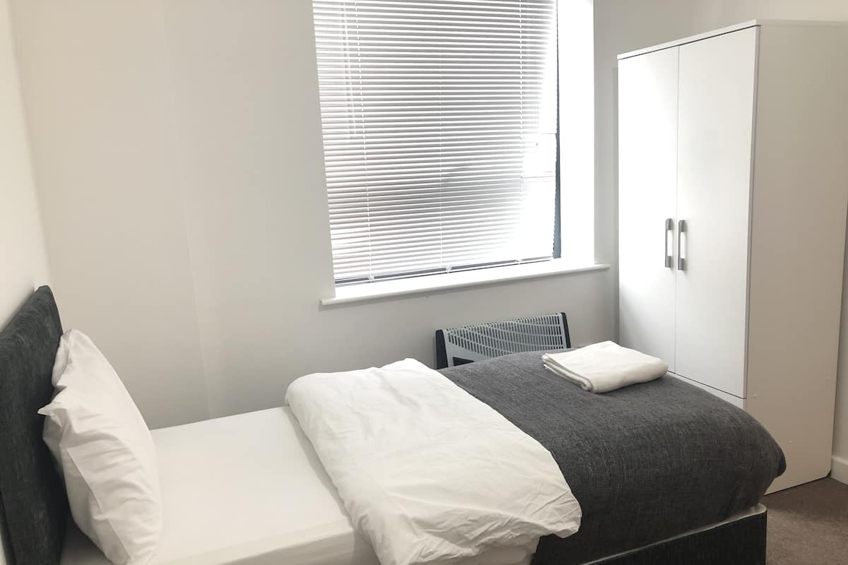 Una cama individual con ropa de cama blanca, una manta gris y una almohada gris. Hay un radiador debajo de la ventana y un armario blanco a la derecha.