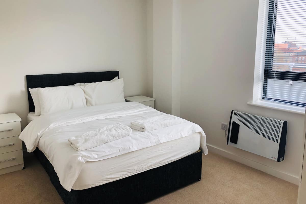 Una cama bien tendida con sábanas blancas en una habitación minimalista con paredes blancas y una ventana con persianas.
