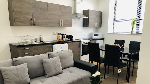 Eine moderne Küche mit dunklen Holzschränken, ein Essbereich mit schwarzen Stühlen und ein graues Sofa mit Kissen.