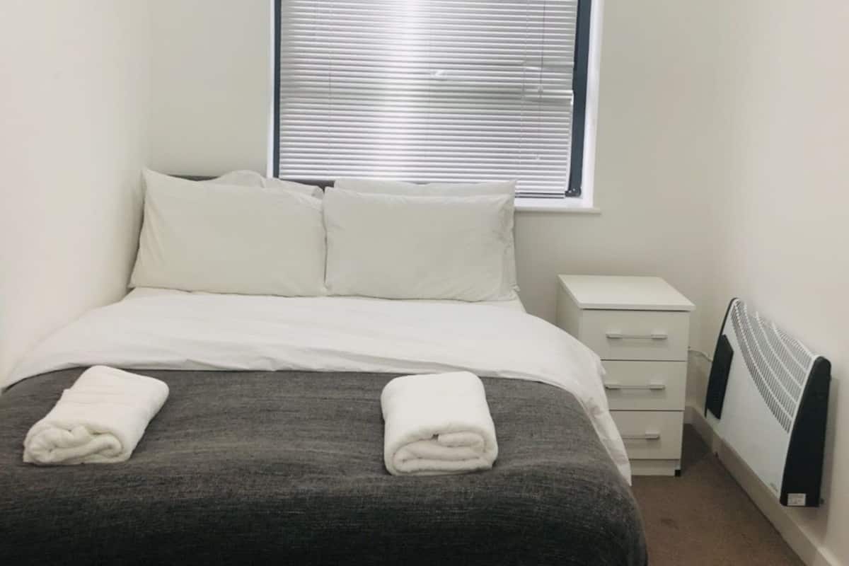 Una habitación pequeña y limpia con cama, mesita de noche, calefacción y ventana con persianas.