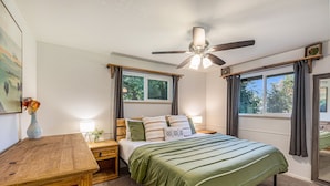 Une chambre à coucher comprenant un lit, des tables de chevet, un bureau en bois, un ventilateur de plafond et des fenêtres avec des rideaux.