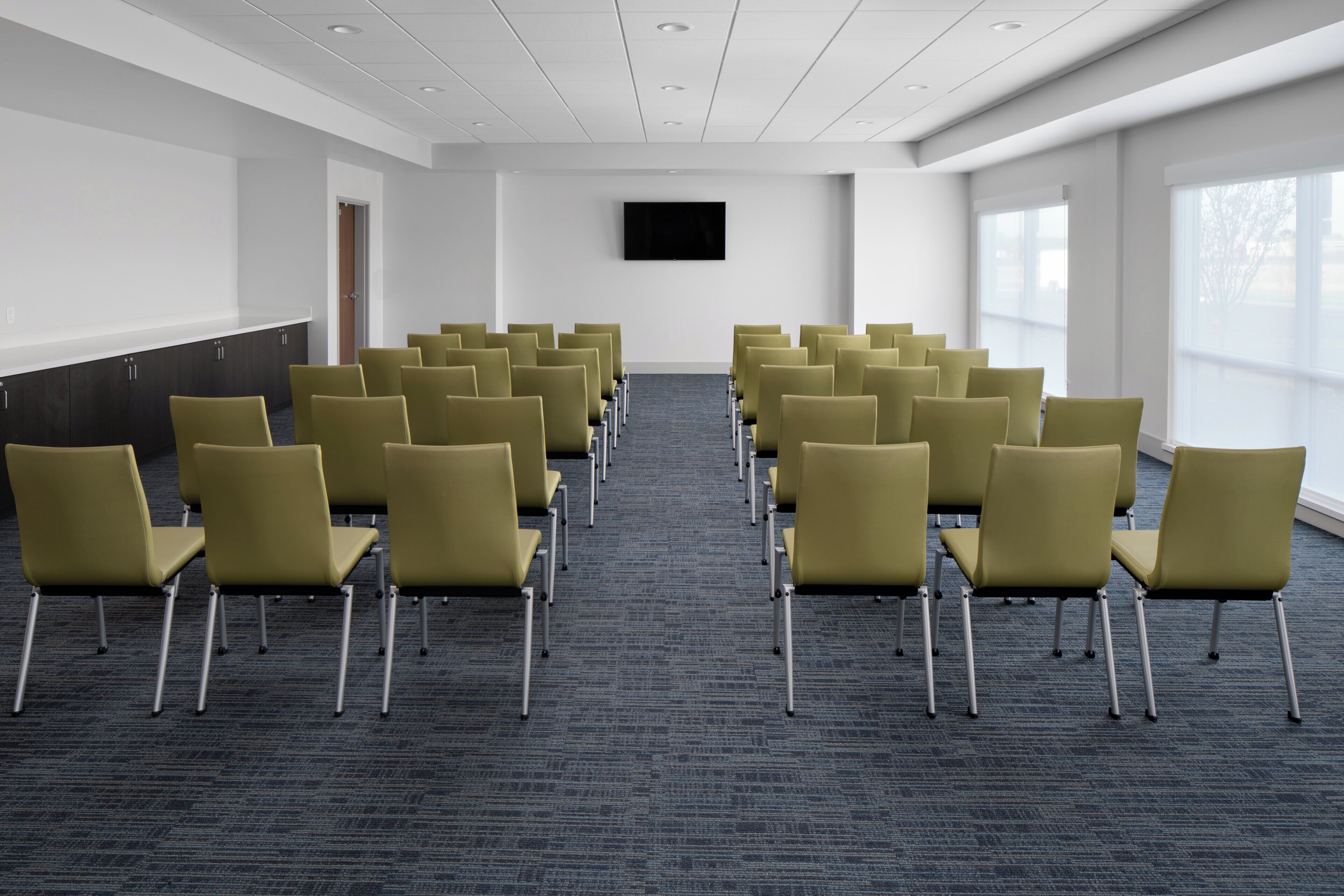 A conference room with rows of chairs, a flat-screen TV on the wall, and large windows on one side.