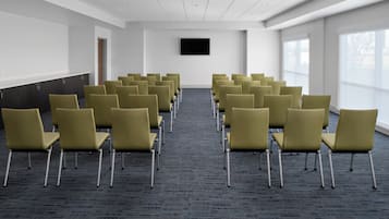 Un salón de conferencias con hileras de sillas, un televisor de pantalla plana en la pared y ventanas amplias a un lado.