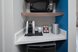 Uma cozinha compacta com cafeteira, xícara de café, caneca e um conjunto de cápsulas de café em uma prateleira branca.