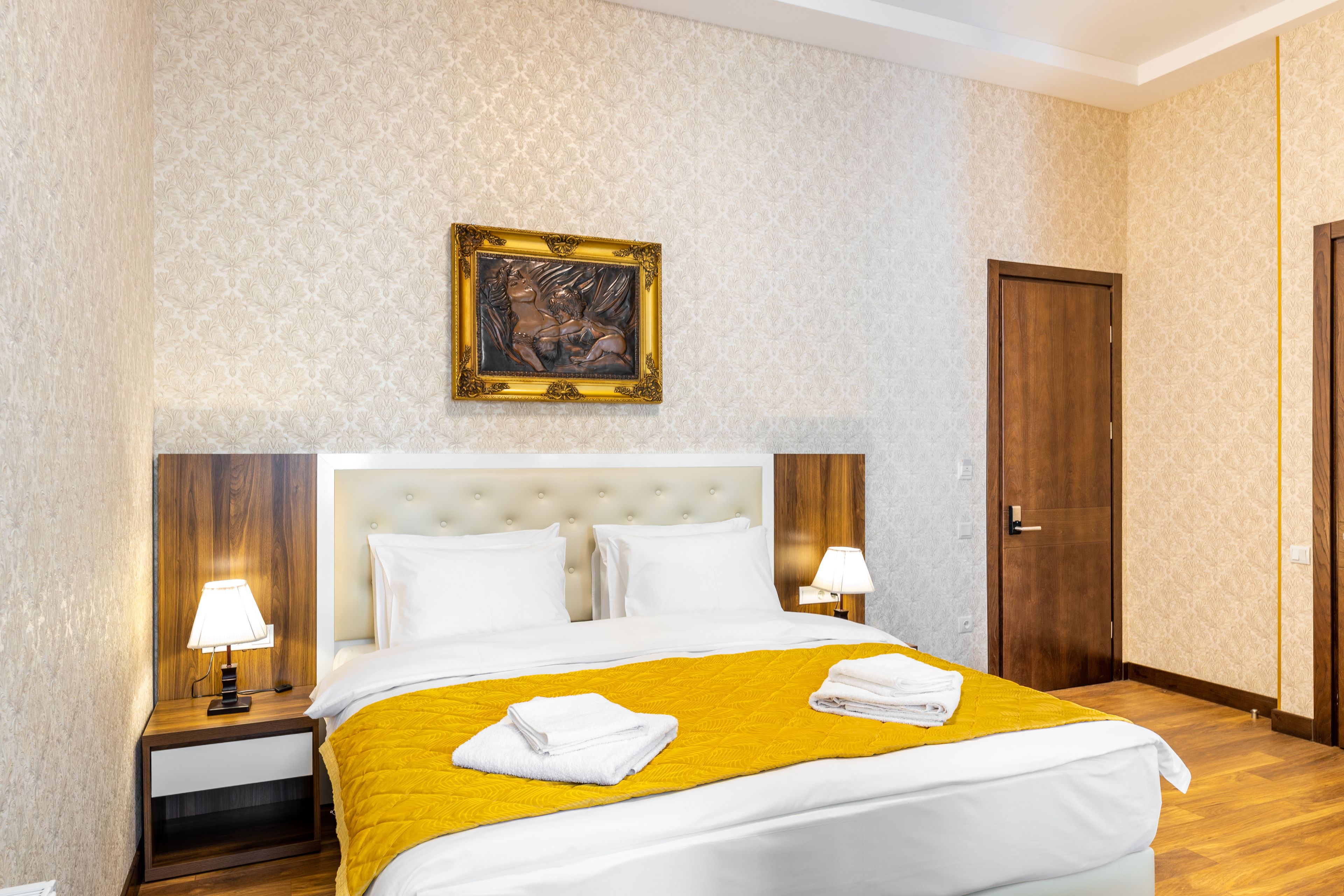Una habitación de hotel con una cama grande, dos mesitas de noche con lámparas, un cabecero de madera, una obra de arte enmarcada en la pared y una puerta de madera.