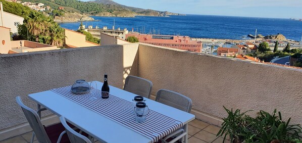 A terrace with a table set for two, overlooking a coastal town with buildings and a view of the sea.