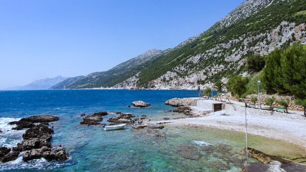 A coastal landscape with clear blue waters, rocky shores, and a mountainous backdrop.