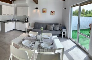A modern kitchen with white cabinets, a dining table set with plates and cutlery, a grey sofa, and a sliding glass door to a green outdoor area. - Domki Magnolia (Kolobrzeg)