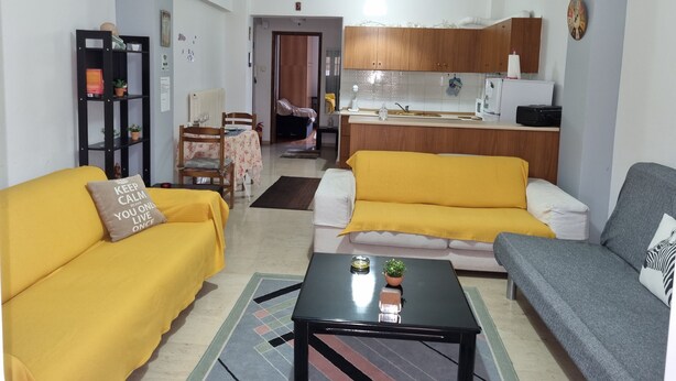 A living room with a yellow sofa, a grey sofa, and a black coffee table. There is a kitchen in the background with wooden cabinets and a sink. - Marie-Claire's Home , Sykies Thessaloniki (Sikies)