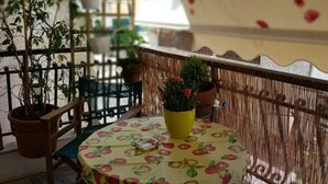 A patio area with a round table covered by a tablecloth with a fruit pattern, surrounded by chairs. There are potted plants on the table and along the railing. - Marie-Claire's Home , Sykies Thessaloniki (Sikies)