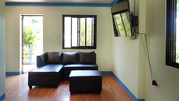 Ein Zimmer mit einer Eckcouch, einem kleinen Hocker, einem an der Wand montierten Fernseher und einem Fenster mit Jalousien.