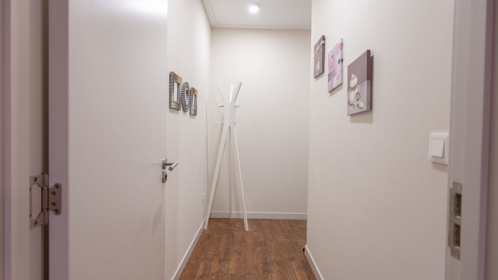 A narrow hallway with white walls and wooden flooring, featuring a door with a metallic handle and a light switch on the wall.