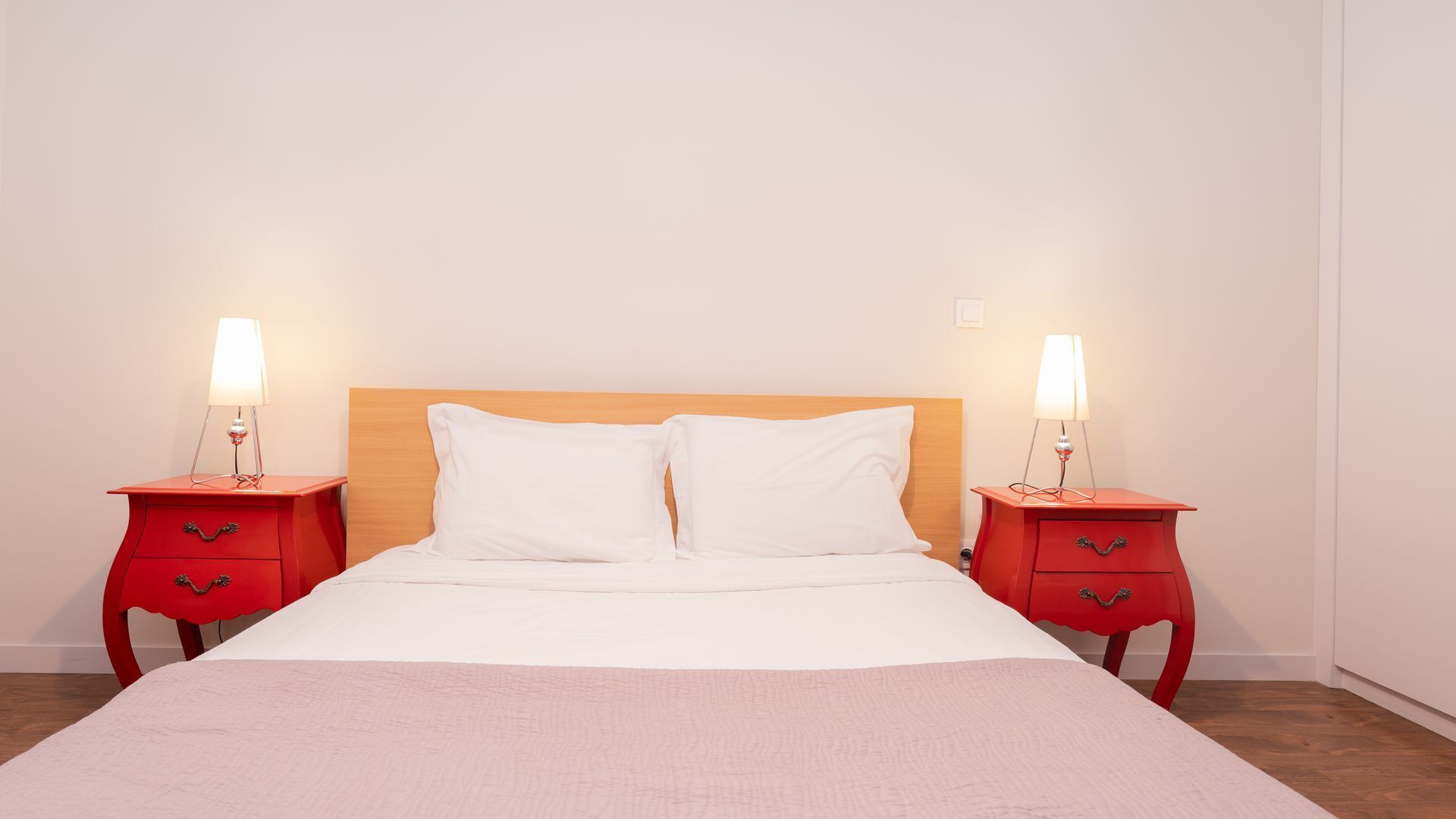 A bed with two bedside tables, each with a lamp, in a room with a plain wall and wooden floor.