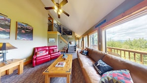 A spacious living room with a red leather sofa, a brown leather sofa, and a wooden coffee table. There is a staircase leading to an upper level, and large windows offering a view of the outdoors. - Views | WiFi | Fireplace | Elk Ridge Lodge (Angel Fire)