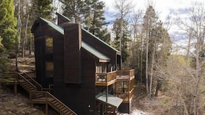 A two-story cabin with a balcony, surrounded by trees and a rocky landscape. - Views | WiFi | Fireplace | Elk Ridge Lodge (Angel Fire)