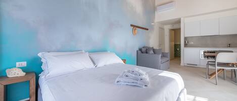 Un dormitorio moderno con una cama grande, una mesita de noche y un sofá gris. Hay una zona de cocina con una mesa de comedor y sillas.