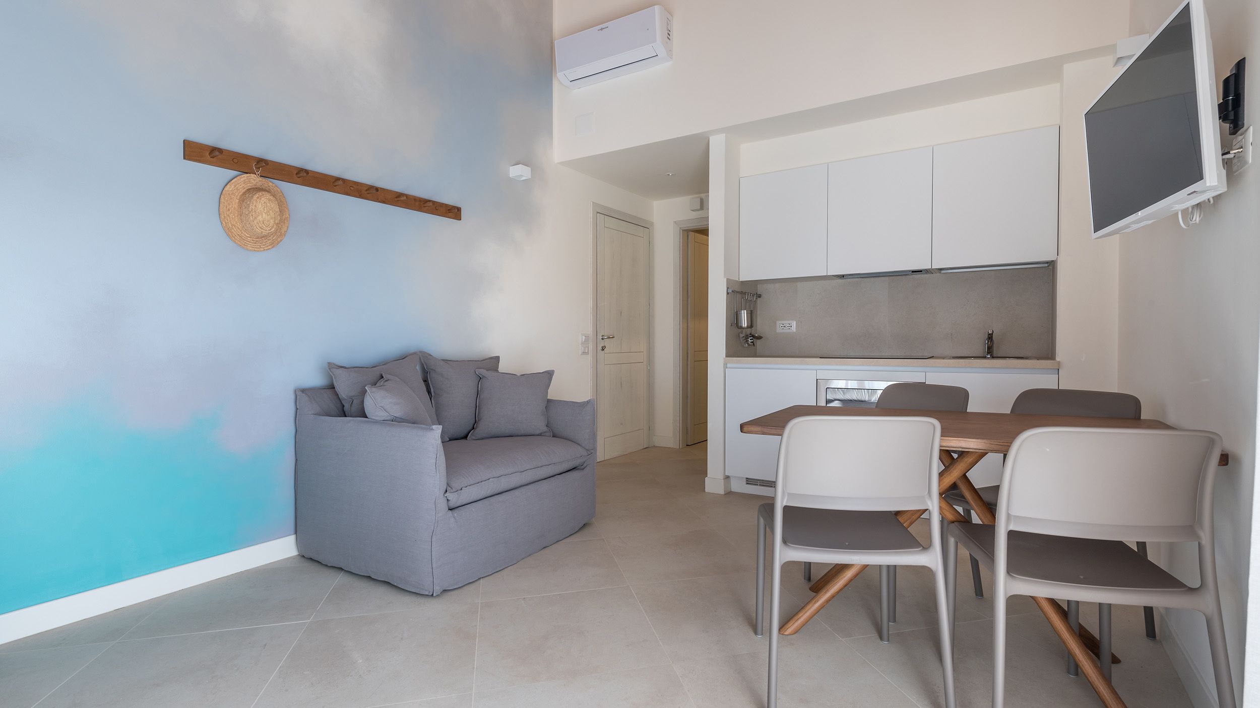 A modern living room with a grey sofa, a dining table with chairs, and a kitchen area with cabinets and a TV.