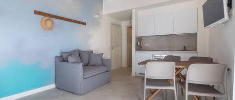 A modern living room with a grey sofa, a dining table with chairs, and a kitchen area with cabinets and a TV.