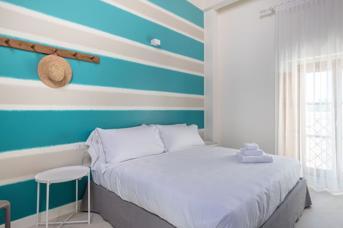 A bedroom with a bed, white bedding, a small bedside table, a wall-mounted hat rack, and a window with sheer curtains.