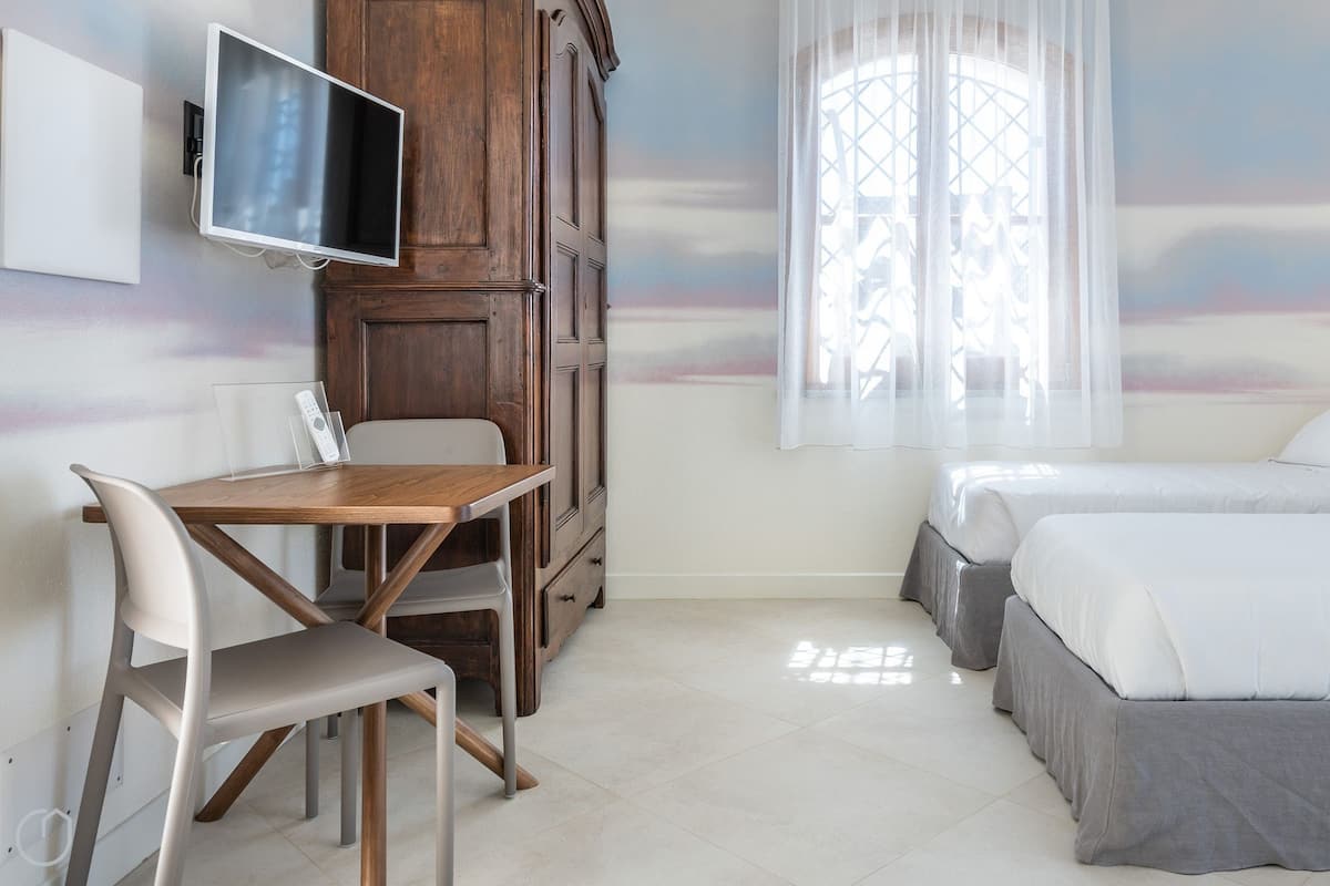 A hotel room with a wooden desk, two beds, a TV, and a large window with sheer curtains.