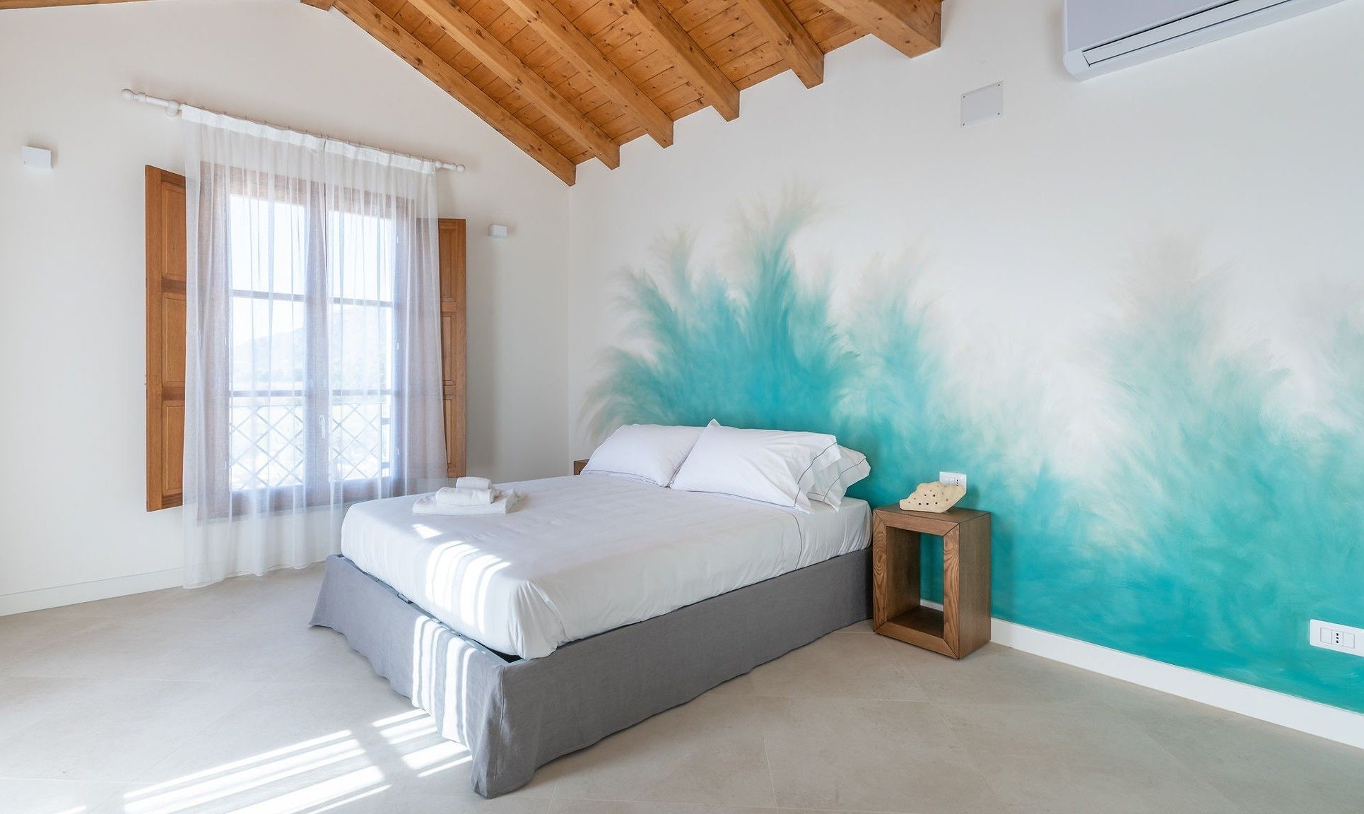 A bedroom with a bed, a nightstand, a window with curtains, and a wall with a painted design.