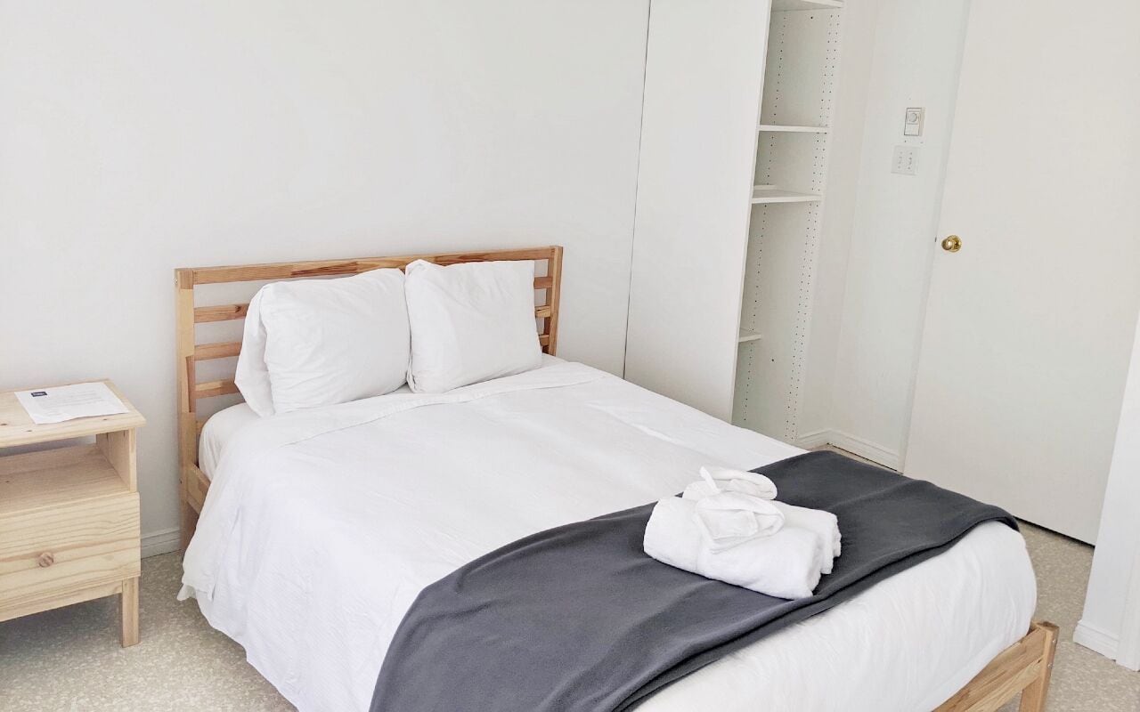 A neatly made bed with white bedding and a gray blanket, a wooden headboard, a bedside table with a lamp, and a white wardrobe with shelves.