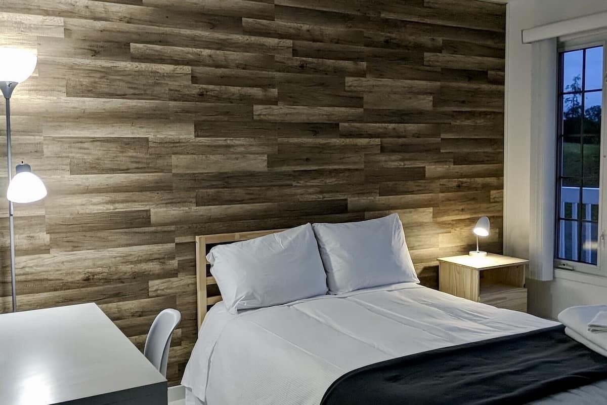 Un dormitorio con pared de madera, cama con ropa de cama blanca, dos lámparas de noche y ventana con vistas a vegetación.