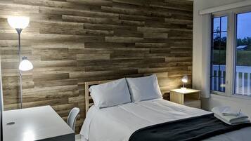Un dormitorio con pared de madera, cama con ropa de cama blanca, dos lámparas de noche y ventana con vistas a vegetación.
