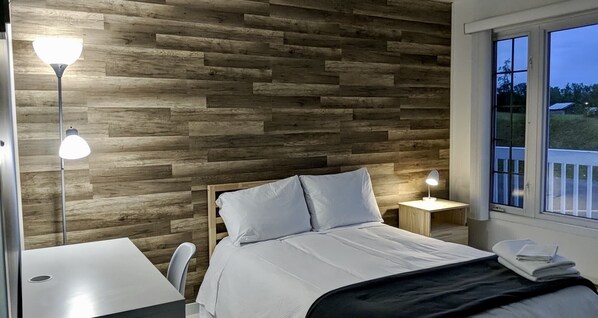 A bedroom with a wooden wall, a bed with white bedding, two bedside lamps, and a window with a view of greenery.