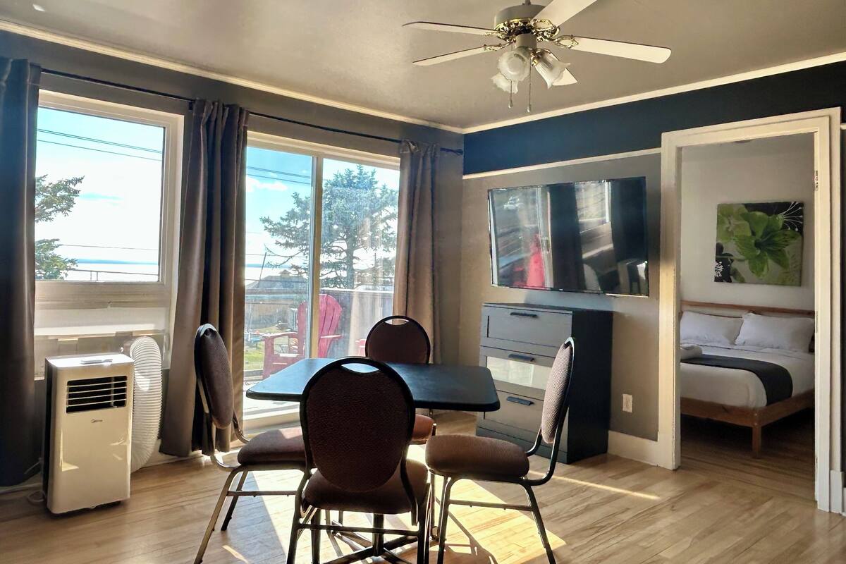 Una habitación con ventilador de techo, una mesa de comedor con sillas, una cama, un televisor y un ventanal con cortinas.