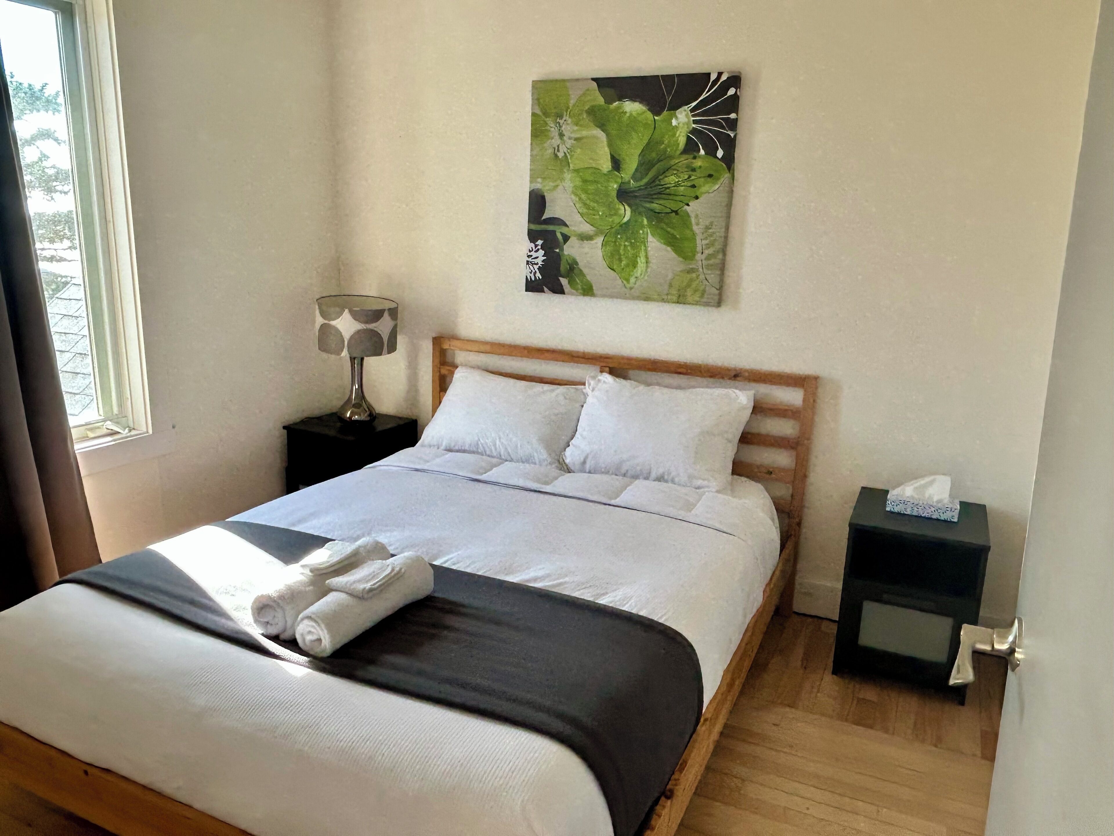 Un lit bien fait, avec du linge de lit blanc et une couverture sombre, une lampe de chevet, une petite table de nuit avec une boîte de mouchoirs, et un tableau représentant des feuilles vertes accroché au mur.