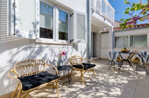 Un patio con sillas de mimbre y una mesa, una ventana con contraventanas y un balcón.
