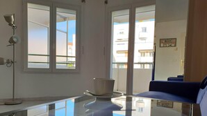 Une pièce moderne avec une table en verre, un canapé bleu et une vue sur les immeubles à travers de grandes fenêtres.