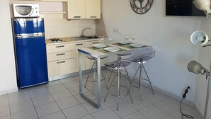 Une cuisine de taille réduite avec un réfrigérateur bleu, une table à manger avec des chaises, une horloge murale et un téléviseur à écran plat.