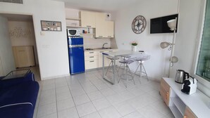 Un ambiente abitativo compatto con angolo cottura, zona pranzo con uno sgabello da bar, divano blu e TV a parete.