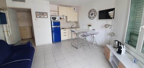 A compact living space with a kitchenette, a dining area with a bar stool, a blue sofa, and a TV mounted on the wall.