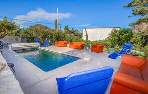 Une piscine bordée de chaises longues orange et bleues, entourée de verdure et d’un mur blanc.