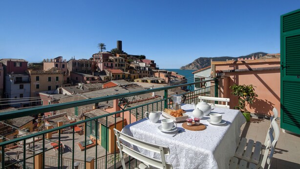 Een balkon met een tafel gedekt voor twee personen, met uitzicht op een kustplaatsje met gebouwen en een helderblauwe lucht.