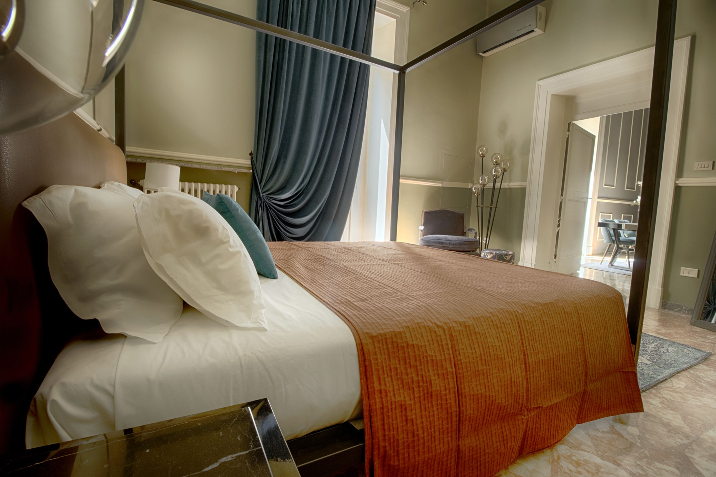 A hotel room with a bed, a nightstand, a window with curtains, a radiator, and a bathroom visible through an open door.