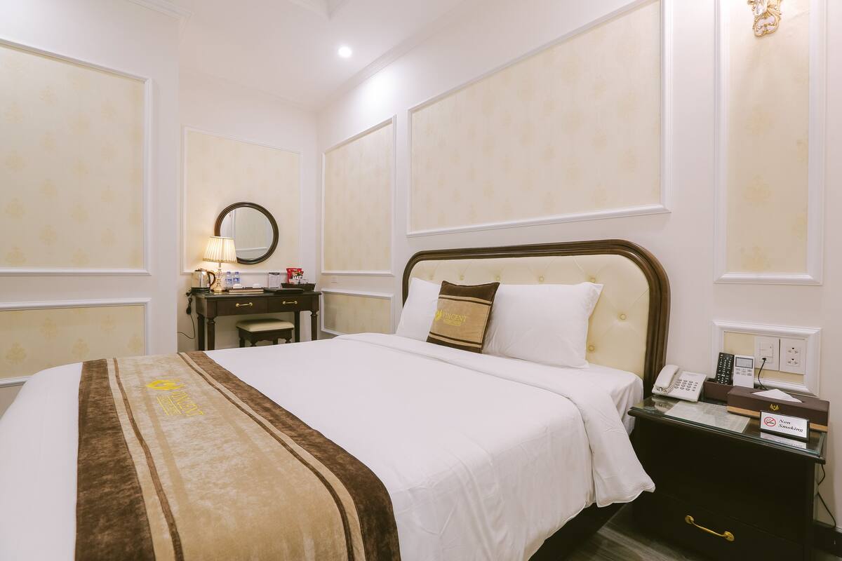 A hotel room with a large bed, a bedside table with a phone and a lamp, a desk with a chair, and a mirror on the wall.