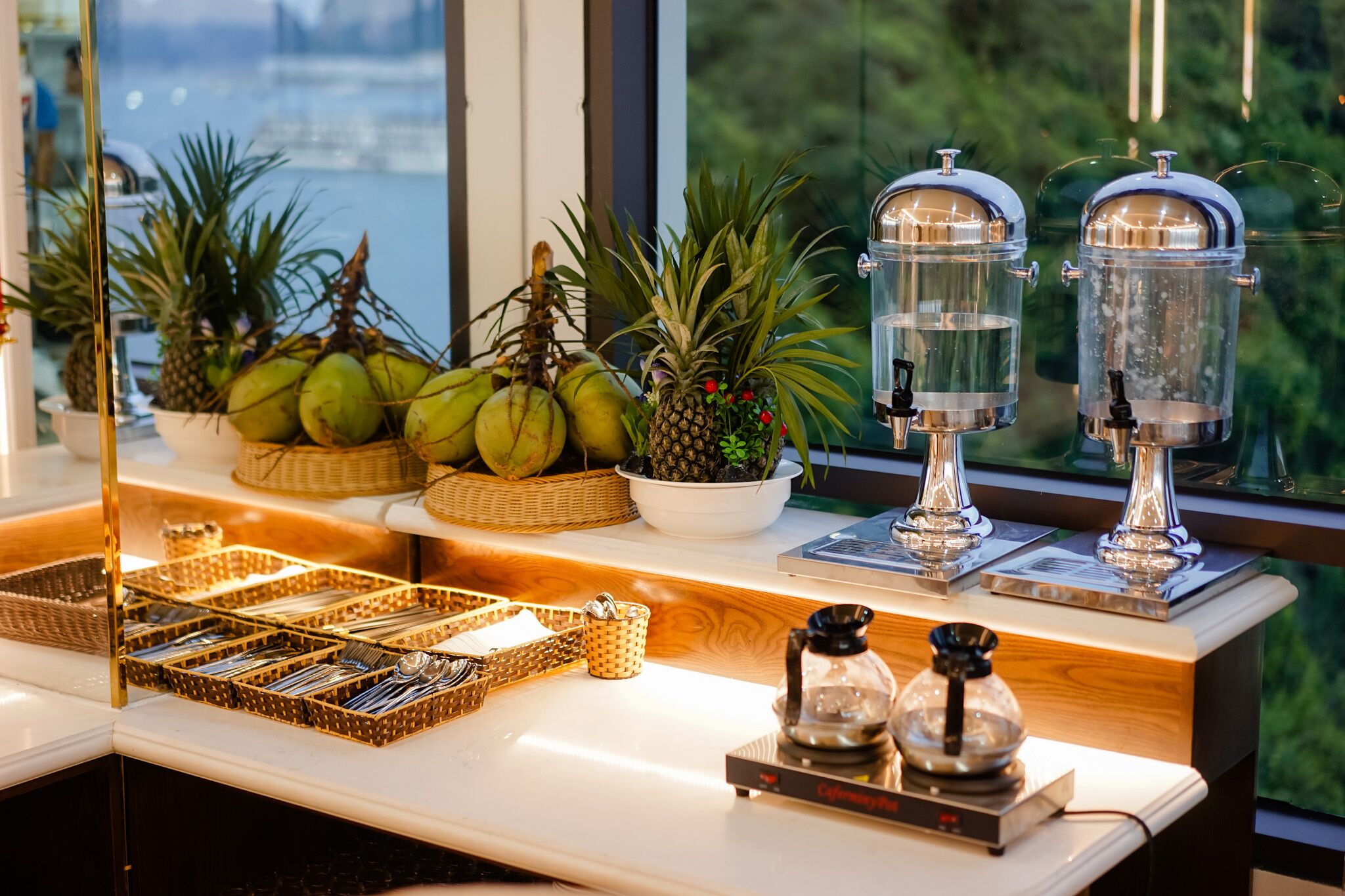 Un bar à petit-déjeuner avec vue sur la ville, comprenant un présentoir de fruits, deux distributeurs d’eau et une machine à café.