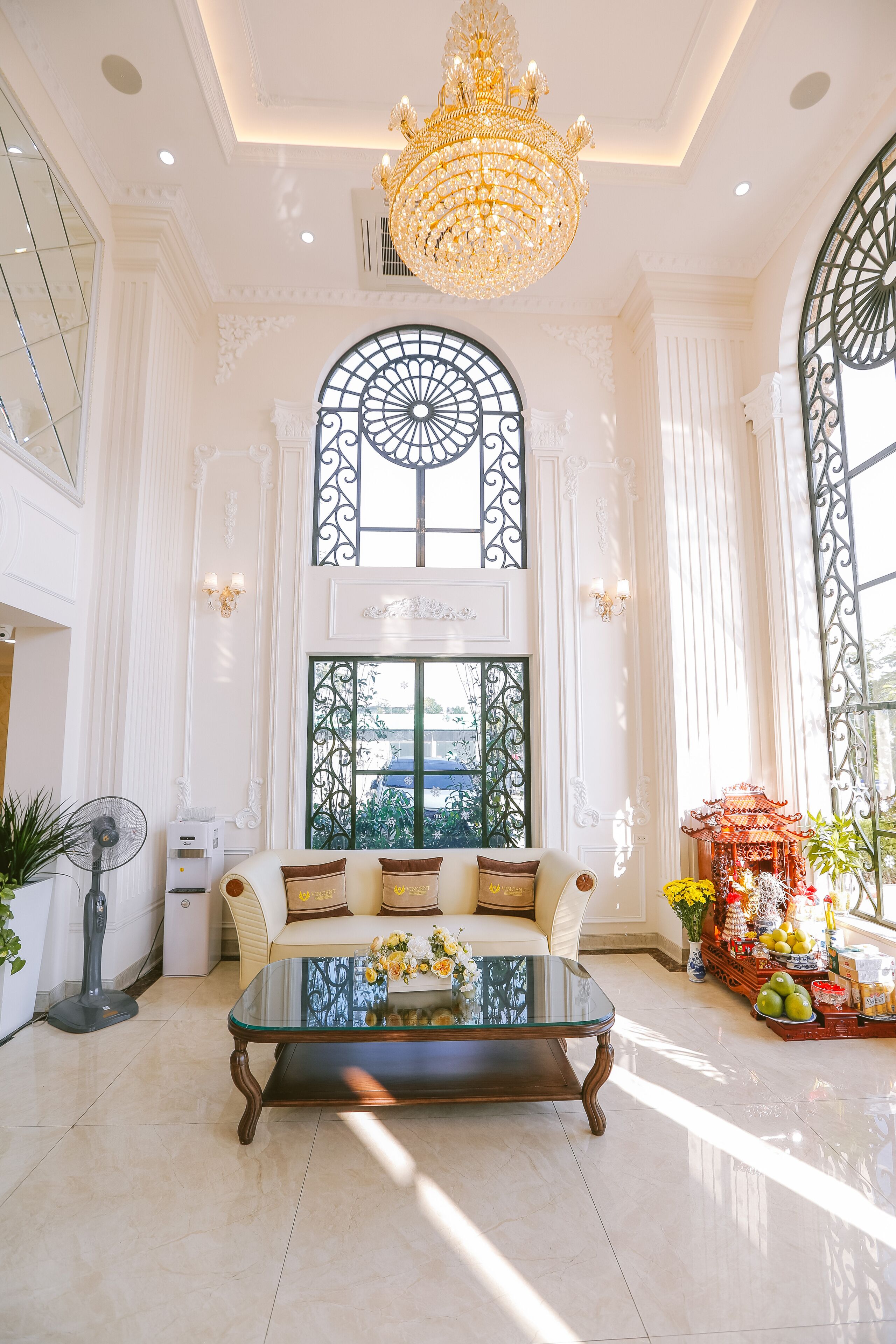 A spacious room with a large glass coffee table, elegant chandelier, and a view through a decorative window.