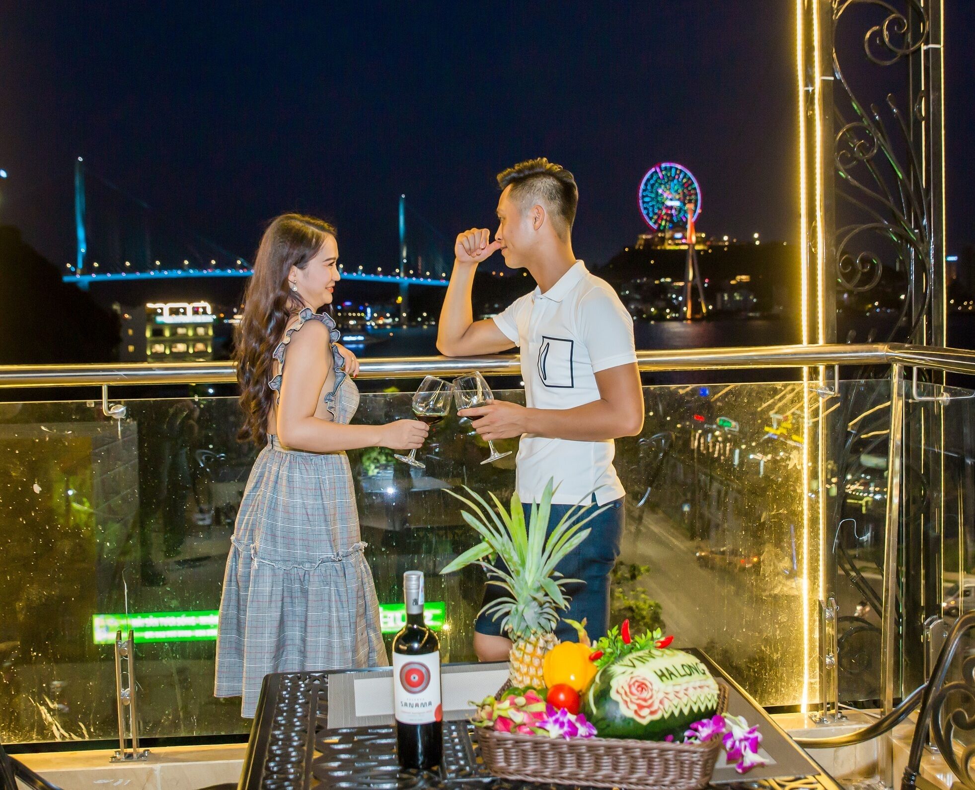 A couple toasting with wine glasses on a balcony with a city view, a bottle of wine, and a fruit basket.