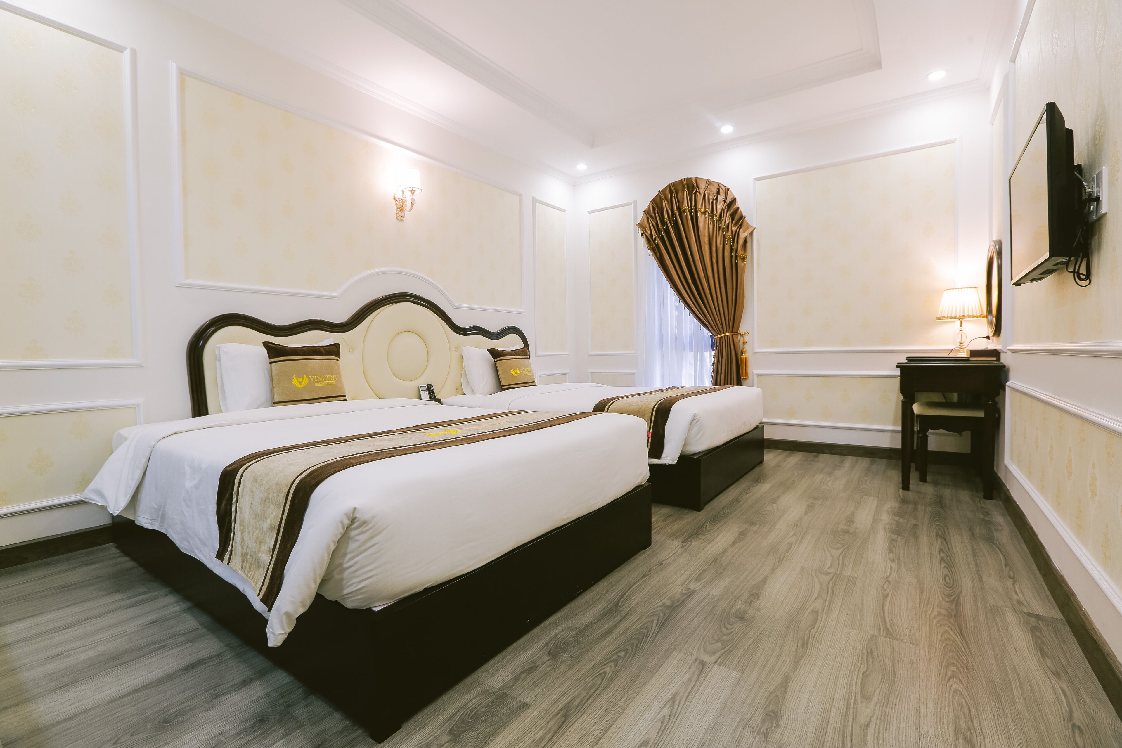 A hotel room with two beds, a nightstand, a lamp, and a large window with curtains.