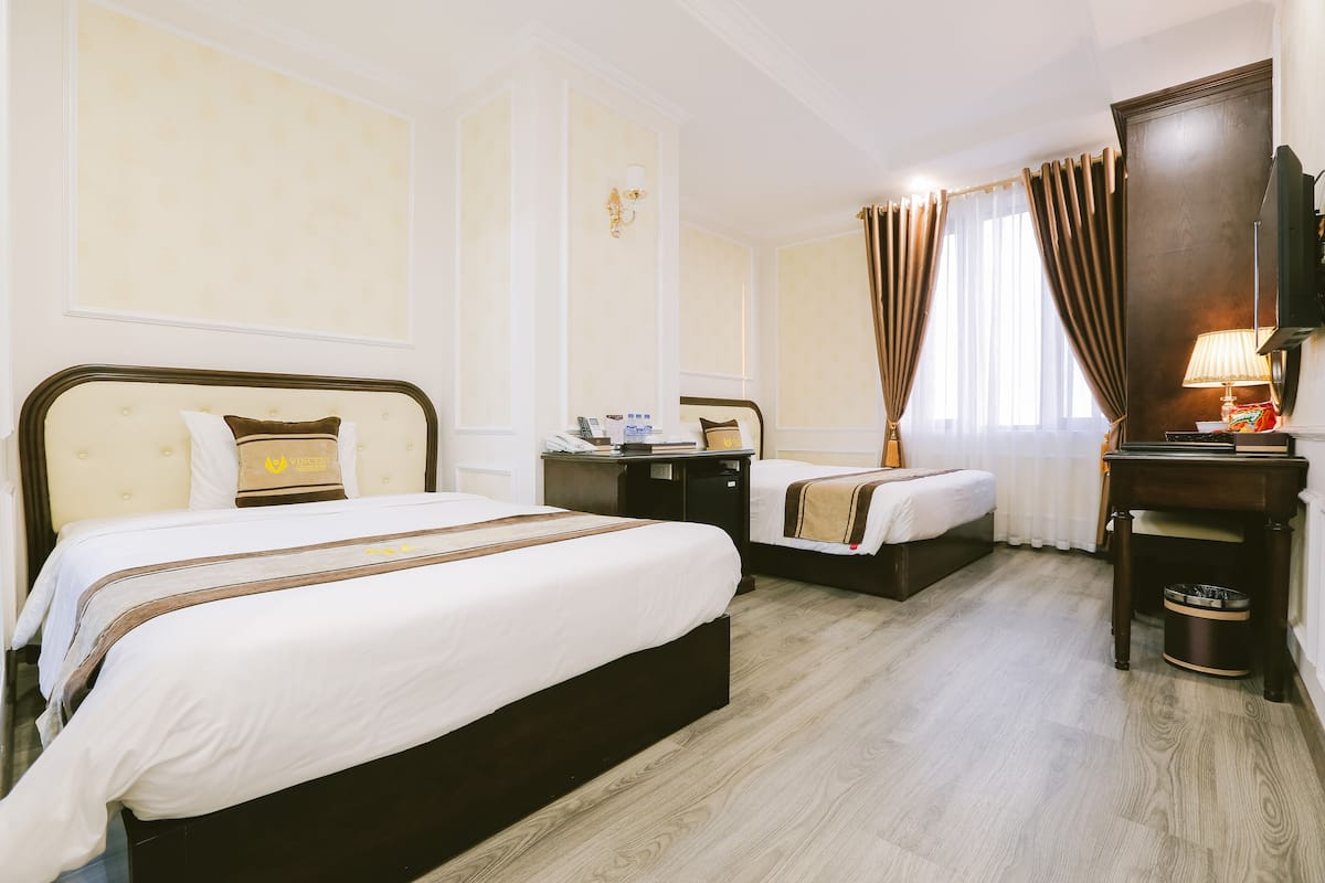 A hotel room with two beds, a wooden floor, a desk with a lamp, and a television.