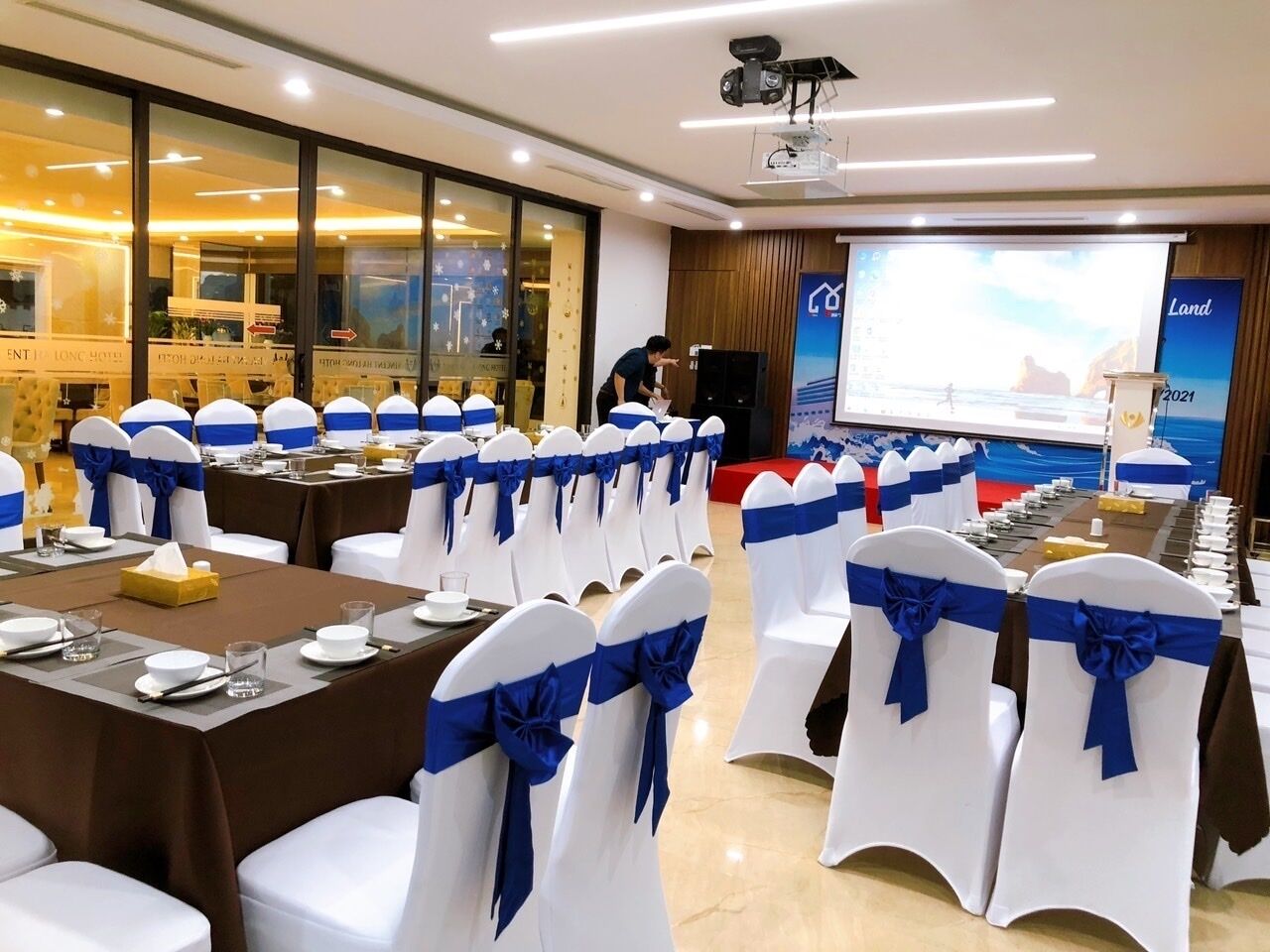 A conference room with rows of white chairs with blue sashes, tables set for a meeting, a projector screen, and a presentation area.
